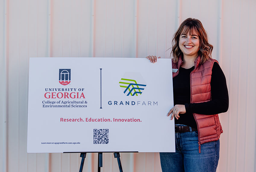A smiling woman with shoulder length hair wearing a red puffer vest, black long-sleeved t-shirt and jeans stands next to a sign celebrating the partnership between UGA and Grand Farm.